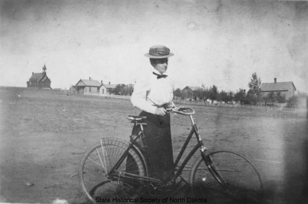 Fannie Dunn Quain standing with her bike. The Bread of Life Church at Camp Hancock State Historic Site is in the background.