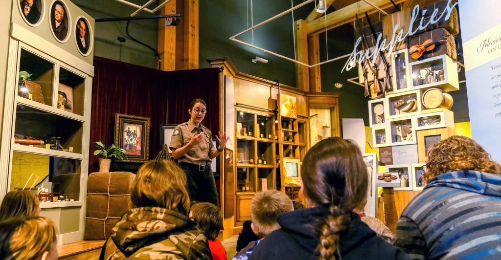 students sit around a female interpreter as she speaks in the middle of a gallery displaying historical arficats