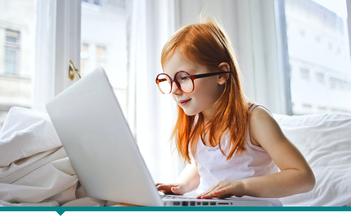 young girl wearing adult glasses sitting on bed with a laptop on her lap