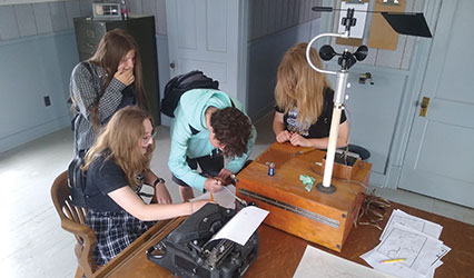 Four youth stand and sit around a wooden table looking and using old weather equipment