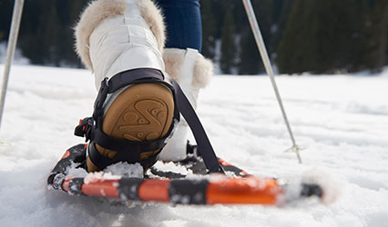 A knee down shot of a woman snowshoeing. She's wearing jeans, white fuzzy boots, and orange snowshoes.