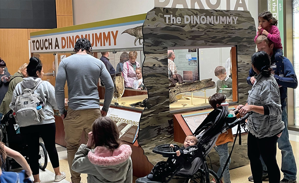 Many people of all ages are gathered around an exhibit case titled The Dinomummy
